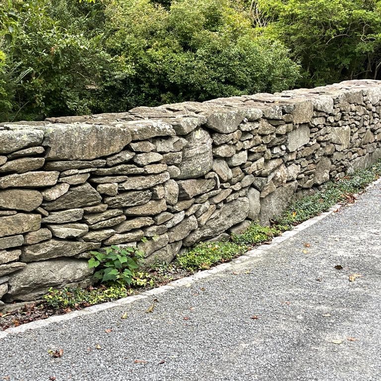 Application image for Fieldstone - New England Flats. The image shows this material being used in a landscape wallstone application. The surface finish shown is weathered. The dominant color shown is gray; with brown, green, white and tan secondaries. The color mix creates a warm feeling. Regular and blotched color patterns.
