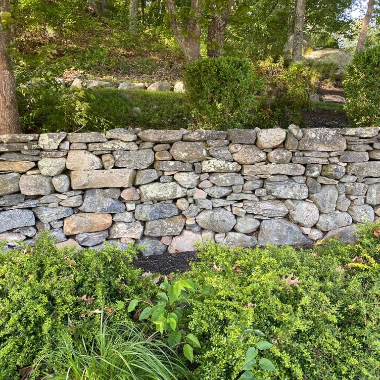 Application image for New England Fieldstone Mix. The image shows this material being used in a landscape wallstone application. The surface finish shown is weathered. The dominant color shown is gray; with beige, brown, white, tan and cream secondaries.