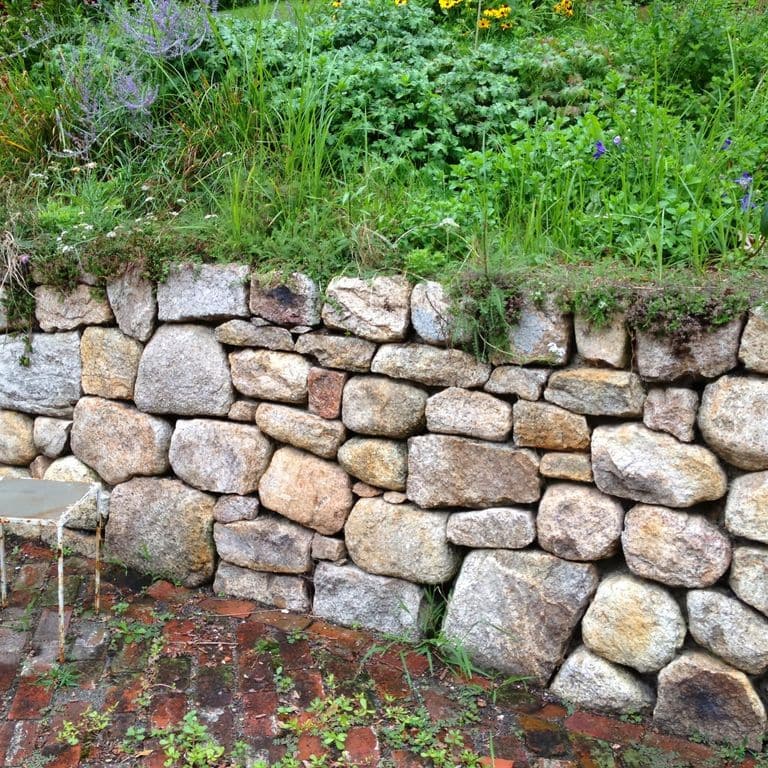 Application image for Fieldstone - New England Mix. The image shows this material being used in a landscape wallstone application. The surface finish shown is weathered. The dominant color shown is gray.