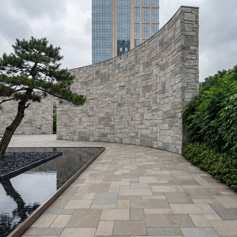 A curved stone wall, featuring exterior veneer (v) made from Stone Curators product M1255 - Nordic Sierra Gray Limestone, stands prominently in an outdoor plaza. A large, shaped pine tree is planted in a bed of dark pebbles next to a reflecting pool, all set against a paved walkway and a backdrop of green bushes and a modern high-rise building under a cloudy sky.