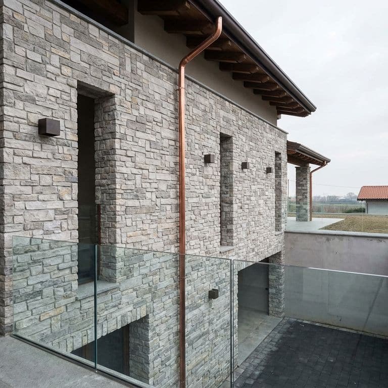 Exterior view of a modern home featuring a prominent exterior veneer (v) is made from Stone Curators product M1255 - Nordic Sierra Gray Limestone. The textured stone facade includes windows, square wall lights, a copper downspout, and a balcony with a glass railing overlooking a lower paved courtyard and garage entrance.