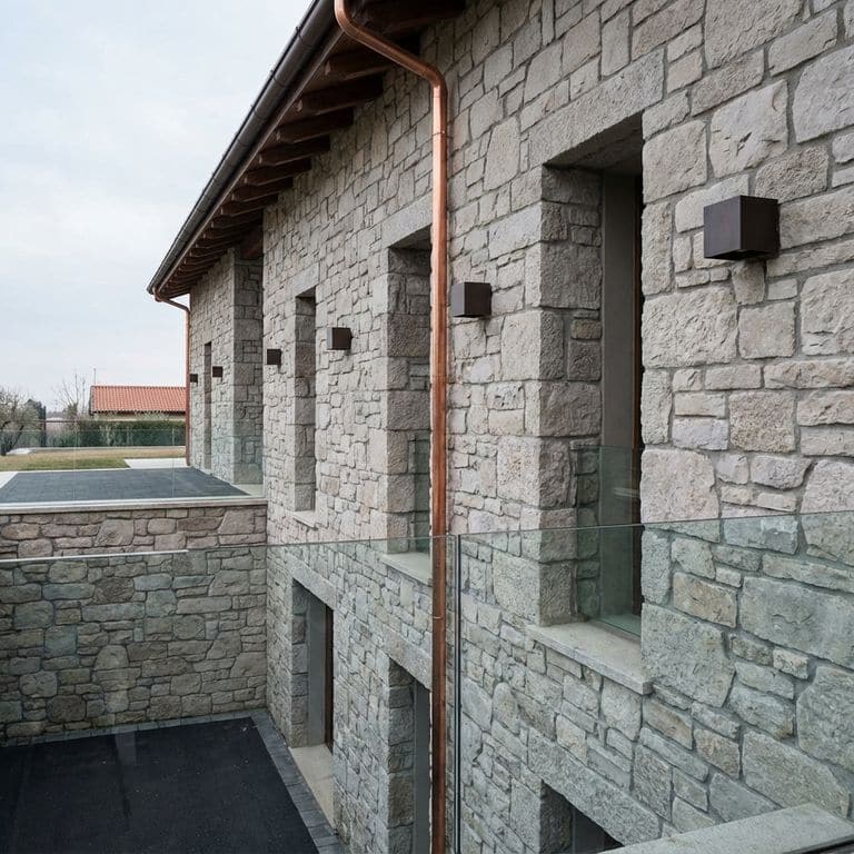 A detailed architectural photo showing a multi-story building featuring a facade of natural grey and blue-hued stone veneer from Stone Curators product M1256 - Fond du Lac Colonial Limestone - Tumbled, along with a prominent vertical copper downspout, modern glass railings, black metal light fixtures, and windows.