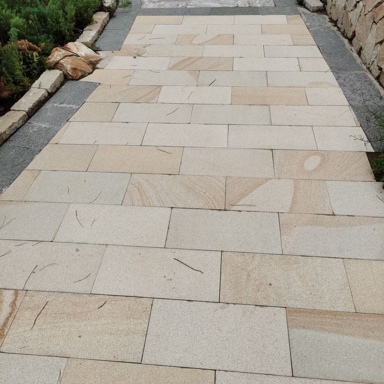 Feature image for Beige Vein Cut Granite. The image shows this material being used in a path pavers application. The surface finish shown is sand blasted. Dominant colors shown are brown and orange/rust. The color mix creates a warm feeling.