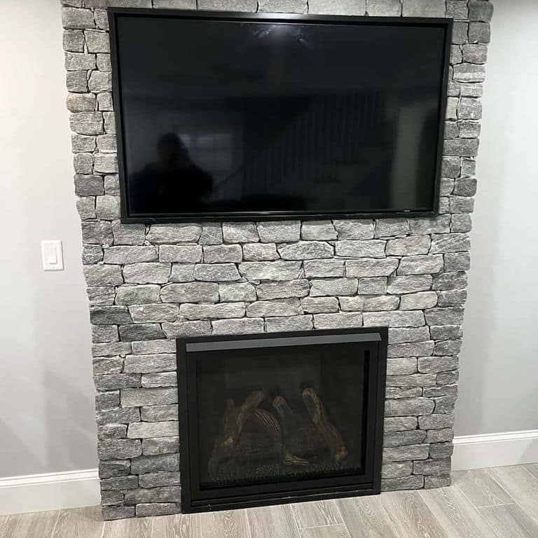 Application image for Oyster Bay Granite. The image shows this material being used for fireplace surround and thin veneer. The surface finish shown is split-face. The dominant color shown is gray. The color mix creates a cool feeling.