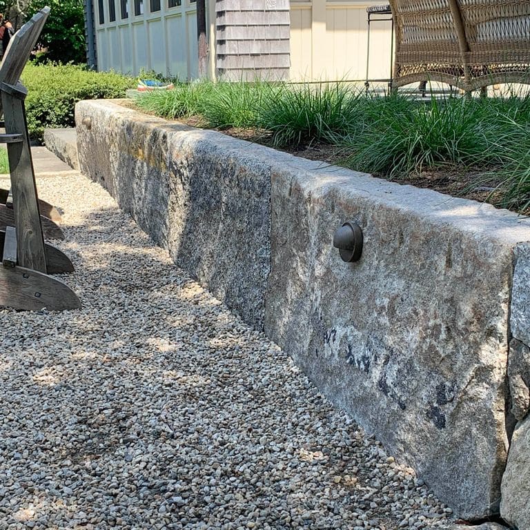 Application image for Reclaimed Granite Curbstone - Jumbo. The image shows this material being used for landscape wallstone and landscape curbing &amp; edging. The surface finish shown is a combination of weathered and worn. The dominant color shown is gray; with a brown secondary. The color mix creates a cool feeling. Speckled, blotched and random color patterns.