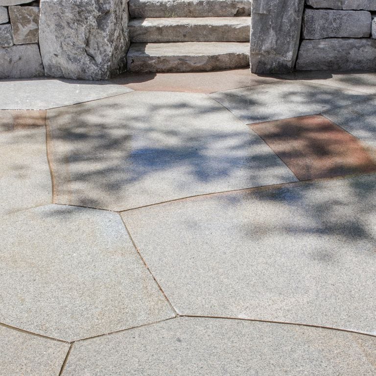 The image shows this material being used for patio pavers and monolithic steps. The dominant color shown is gray; with beige and brown secondaries. Fine speckled color pattern.