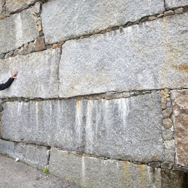 Feature image for Reclaimed Granite Bridge Blocks. The image shows this material being used in a landscape wallstone application. The design pattern shown is dimensional squares &amp; rectangles. The dominant color shown is gray; with beige, brown, orange/rust, white and cream secondaries. The color mix creates a warm feeling.