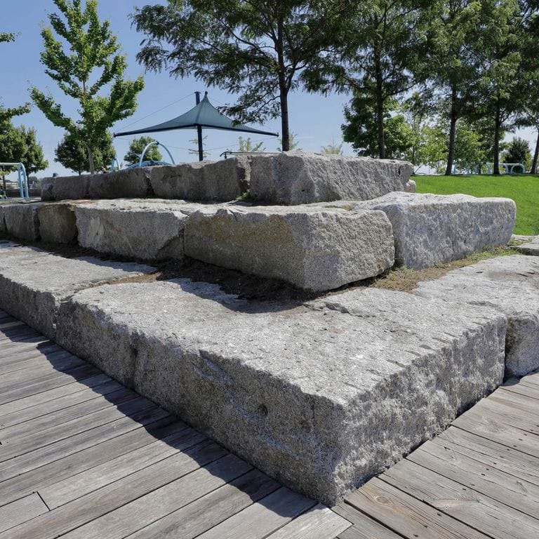 Application image for Reclaimed Granite Bridge Blocks. The image shows this material being used in a seating &amp; benches application. The surface finish shown is a combination of chiseled and weathered. The dominant color shown is gray. The color mix creates a cool feeling.