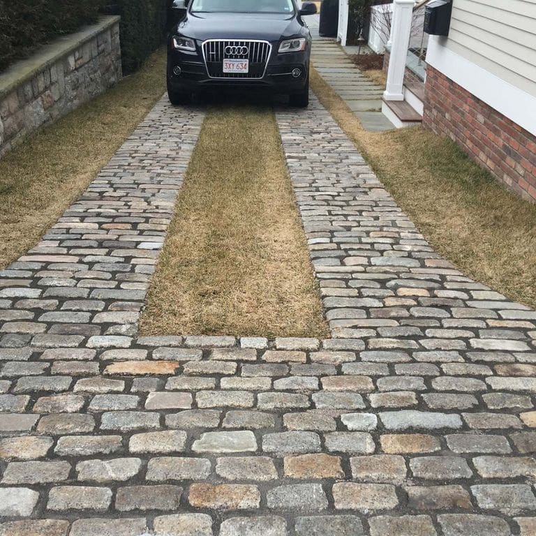 Feature image for Reclaimed New England Granite Cobblestones - Medium. The image shows this material being used in a driveway pavers application. The design pattern shown is running bond with tops up. Dominant colors shown are brown and gray; with beige, green, orange/rust, purple, white and tan secondaries. The color mix creates a warm feeling. Regular and speckled color patterns.