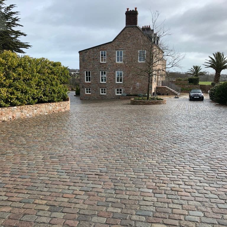 Application image for Reclaimed Swedish Granite Cobblestones. The image shows this material being used in a driveway pavers application. The surface finish shown is antiqued. Dominant colors shown are gray and red. The color mix creates a warm feeling.