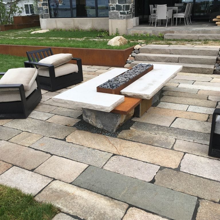 The image shows this material being used in a patio pavers application. The surface finishes shown are weathered and worn. Dominant colors shown are gray and tan; with beige, brown and orange/rust secondaries. The color mix creates a warm feeling. Regular and fine speckled color patterns.