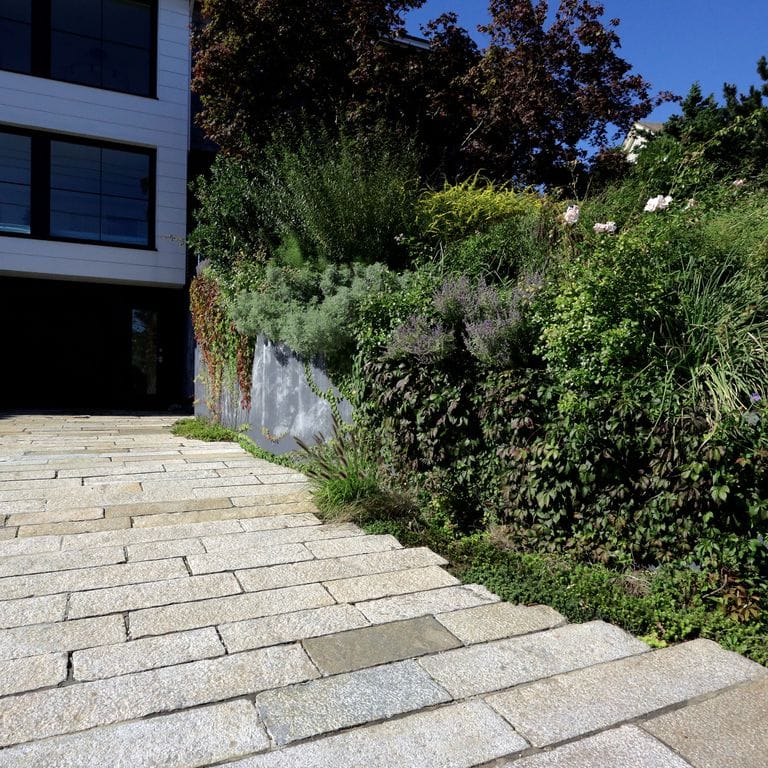 Application image for Reclaimed Footworn Granite Planks. The image shows this material being used in a driveway pavers application. The surface finish shown is a combination of weathered and worn. Dominant colors shown are beige and gray; with pink, purple, white, tan and cream secondaries. The color mix creates a warm feeling. Regular and fine speckled color patterns.