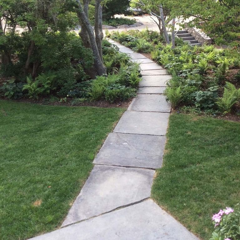 Feature image for Reclaimed Bluestone Sidewalk Stones. The image shows this material being used in a path pavers application. The design pattern shown is roughly square roughly rectangular. The dominant color shown is gray; with blue, purple and white secondaries. The color mix creates a cool feeling. Regular color pattern.