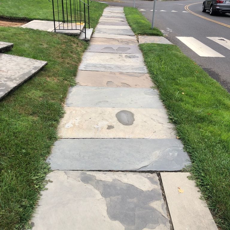 Application image for Reclaimed Bluestone Sidewalk Stones. The image shows this material being used in a path pavers application. The surface finish shown is a combination of natural cleft, weathered and worn. Dominant colors shown are beige, gray and cream; with pink, purple, white and tan secondaries. The color mix creates a warm feeling. Regular and blotched color patterns.