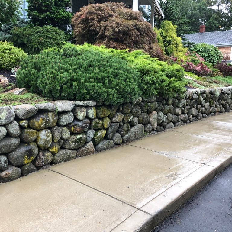 Application image for New England Fieldstone Rounds. The image shows this material being used in a landscape wallstone application. The surface finish shown is weathered. The dominant color shown is gray.