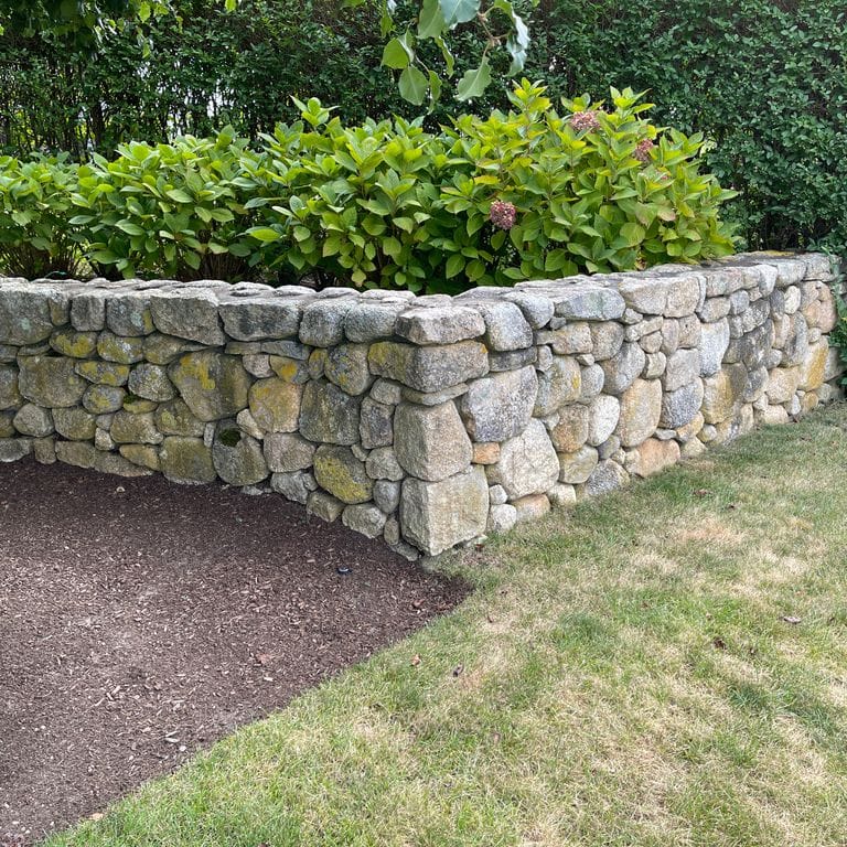 Application image for New England Fieldstone Rounds. The image shows this material being used in a landscape wallstone application. The surface finish shown is weathered. Dominant colors shown are gray and tan; with brown, orange/rust and white secondaries. The color mix creates a warm feeling. Speckled color pattern.