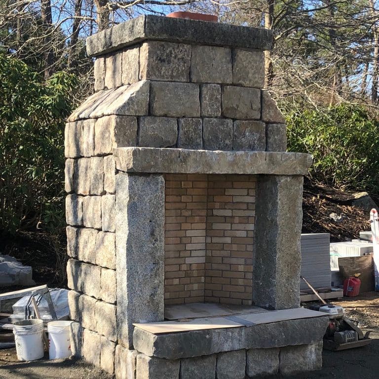 Application image for Reclaimed New England Granite Cobblestones - Road Pavers. The image shows this material being used for fireplace surround and outdoor fire concepts. The surface finish shown is a combination of weathered and worn. Dominant colors shown are beige and tan; with brown, gray, orange/rust, white, yellow and cream secondaries.
