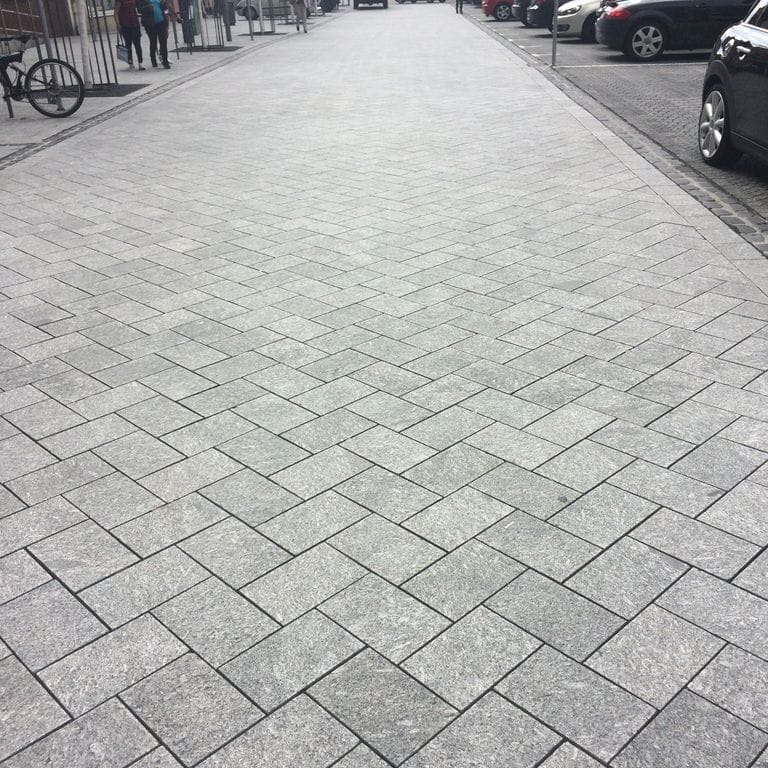 Feature image for Granite - Colonial Blue. The image shows this material being used for driveway pavers and public area. The dominant color shown is gray; with beige, black, pink, purple and white secondaries. Exhibits "movement" and the color mix creates a cool feeling. Swirled, blotched and coarse speckled color patterns. Contains mineral inclusions.