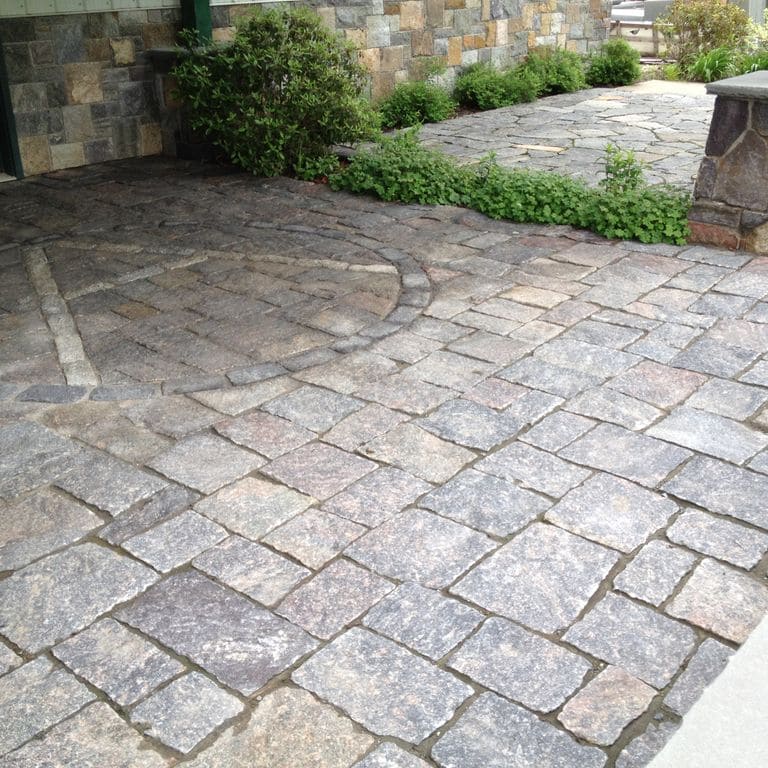 Application image for Adirondack Granite. The image shows this material being used in a patio pavers application. The surface finish shown is split-face with flat plane. Dominant colors shown are gray and white; with beige, blue, purple and cream secondaries. The color mix creates a cool feeling. Regular and fine speckled color patterns.