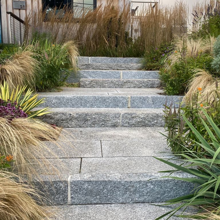 Feature image for Freshwater Pearl Granite. The image shows this material being used in a monolithic steps application. The dominant color shown is gray; with beige, black, pink, purple, white, tan and cream secondaries. The color mix creates a cool feeling. Speckled, coarse speckled and blotched color patterns. Contains some mica and mineral inclusions.