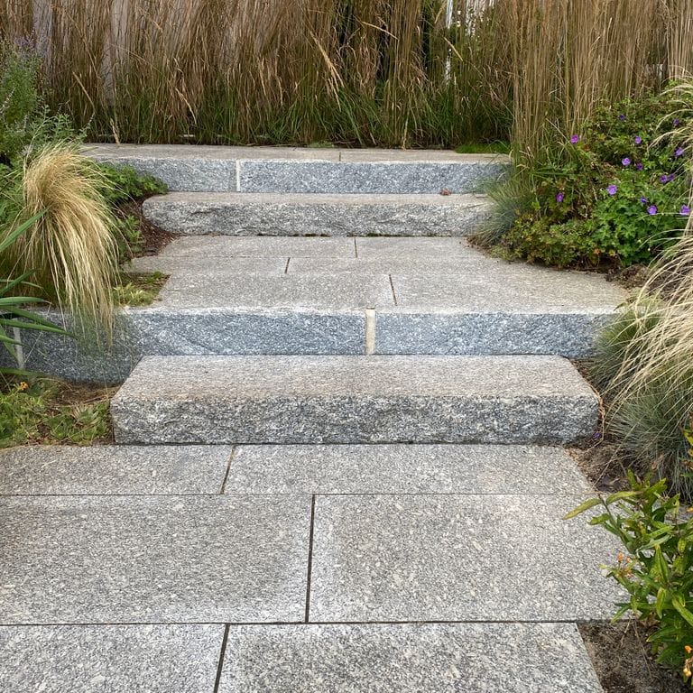 Application image for Freshwater Pearl Granite. The image shows this material being used for path pavers and monolithic steps. The surface finish shown is flamed. The dominant color shown is gray. The color mix creates a warm feeling. Speckled color pattern.
