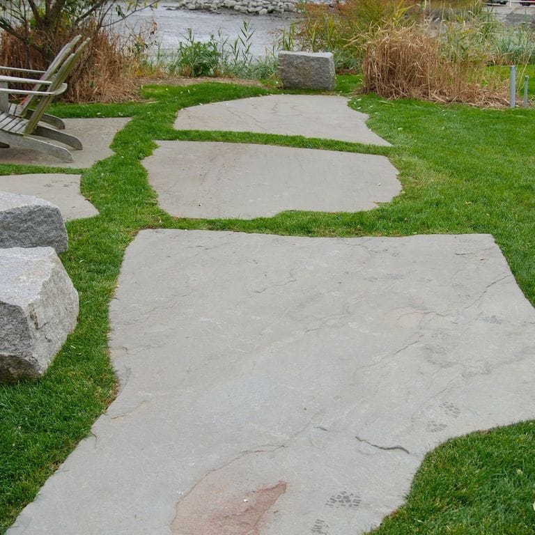 Feature image for Bluestone Natural Cleft - Green/Brown. The image shows this material being used in a path pavers application. The dominant color shown is gray; with beige and white secondaries. The color mix creates a cool feeling. Cloudy color pattern.