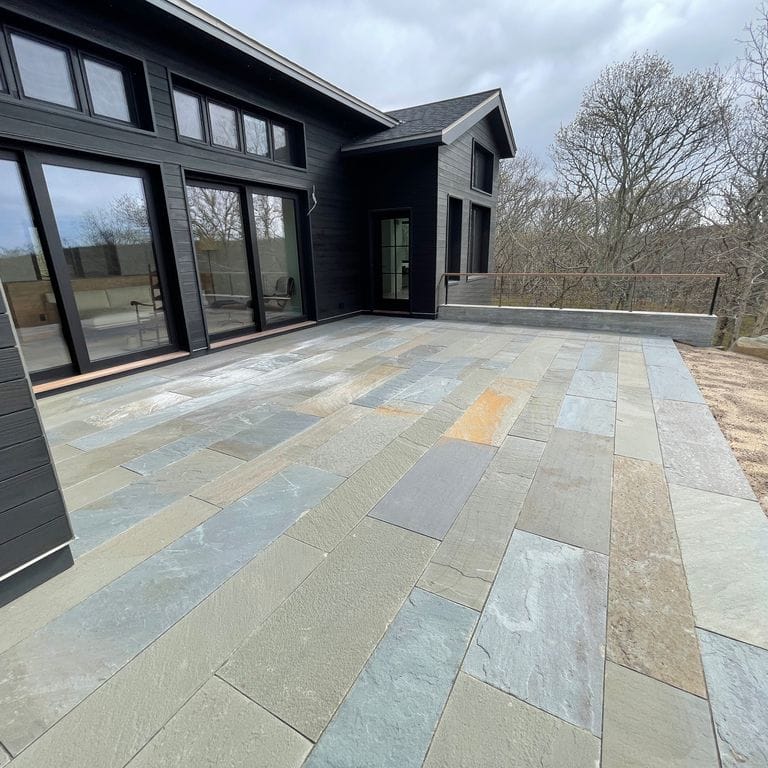 Feature image for Full Color Mix Bluestone - Natural Cleft. The image shows this material being used in a patio pavers application. The surface finish shown is natural cleft. Dominant colors shown are blue and gray; with blue and orange/rust secondaries. The color mix creates a cool feeling. Regular and random color patterns.