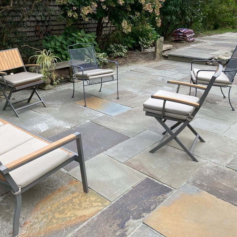 Application image for Bluestone Natural Cleft - Gunmetal. The image shows this material being used in a patio pavers application. The surface finish shown is natural cleft. Dominant colors shown are brown, green and gray; with an orange/rust secondary.