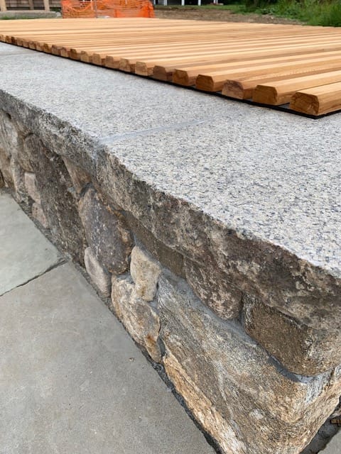 Application image for Reclaimed Rockport Granite. The image shows this material being used in a wallstone cap application. This is a building and landscape stone for indoor and outdoor commercial and residential applications. Suitable for general use, freeze-thaw conditions, low heat absorption surfaces, high traffic areas, low slipperiness surfaces and high heel compatibilty situations. Moderate to high on the price spectrum.
