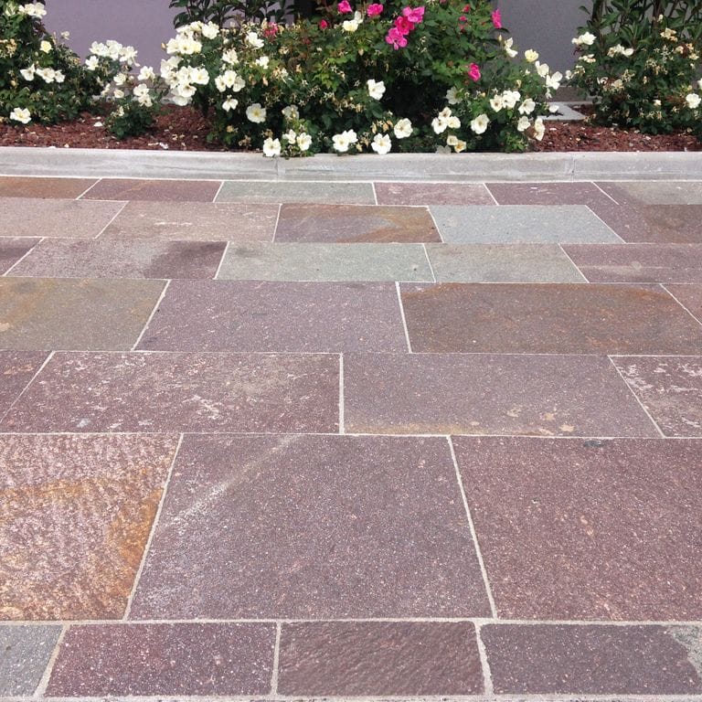 Feature image for Porphyry - Natural Surface - Mixed Red. The image shows this material being used in a patio pavers application. Dominant colors shown are gray, orange/rust and purple; with beige, brown, green, pink and tan secondaries. Exhibits "movement" and the color mix creates a warm feeling. Speckled and random color patterns.