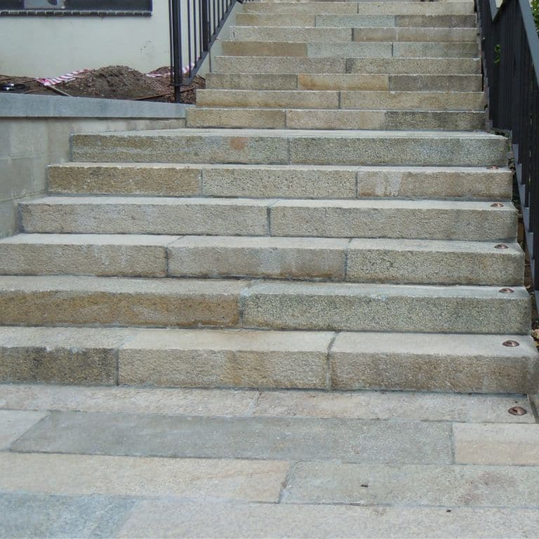 Feature image for Reclaimed Granite Stair Blocks. The image shows this material being used in a monolithic steps application. The dominant color shown is gray; with beige, blue, brown, green, orange/rust, purple, white and tan secondaries. Exhibits "movement". Speckled and blotched color patterns.