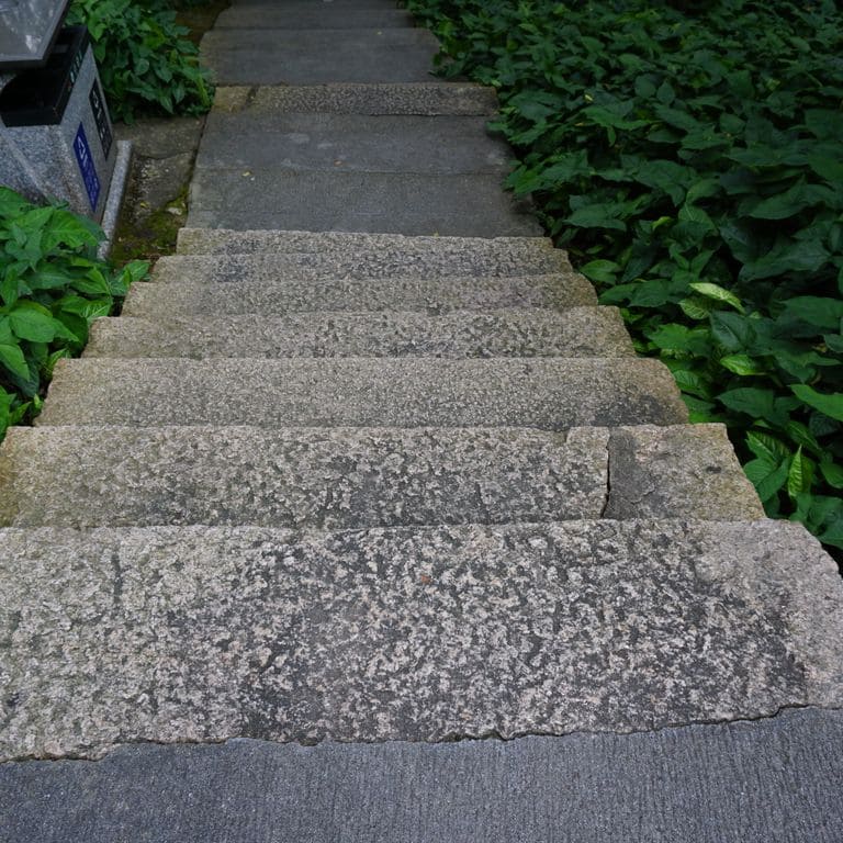 Application image for Reclaimed Granite Stair Blocks. The image shows this material being used in a monolithic steps application. The surface finish shown is a combination of weathered and worn. The dominant color shown is gray; with blue, brown, green, purple and white secondaries. The color mix creates a cool feeling. Speckled and blotched color patterns.