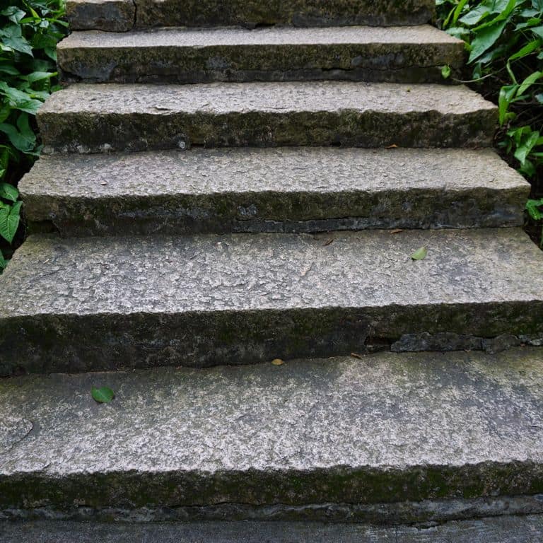 Application image for Reclaimed Granite Stair Blocks. The image shows this material being used in a monolithic steps application. The surface finish shown is a combination of weathered and worn. The dominant color shown is gray; with blue, green, purple and white secondaries. The color mix creates a cool feeling.