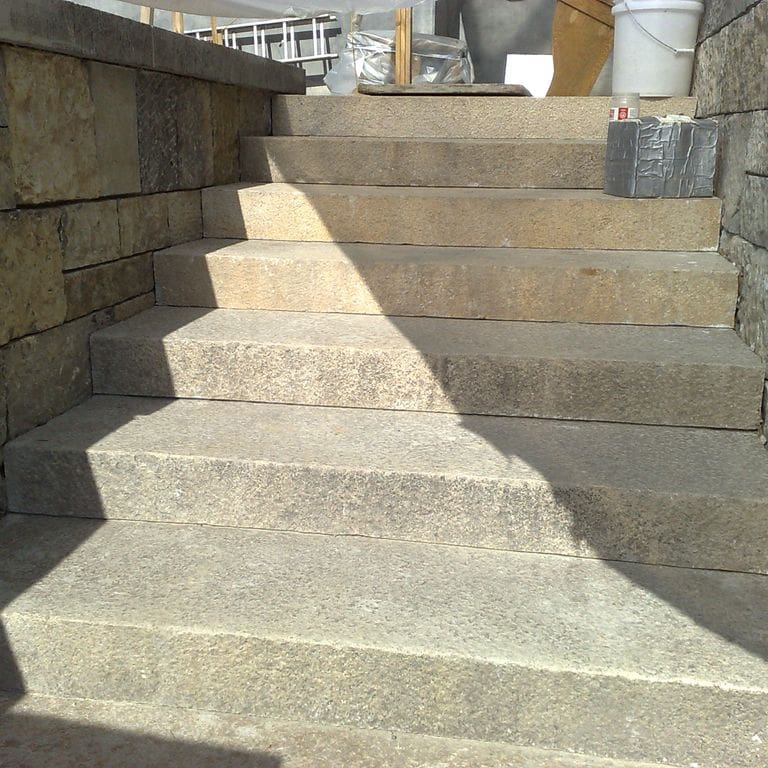 Application image for Reclaimed Granite Stair Blocks. The image shows this material being used in a monolithic steps application. The surface finish shown is a combination of weathered and worn. Dominant colors shown are beige and tan; with brown, gray, orange/rust, white and cream secondaries. The color mix creates a warm feeling. Speckled color pattern.