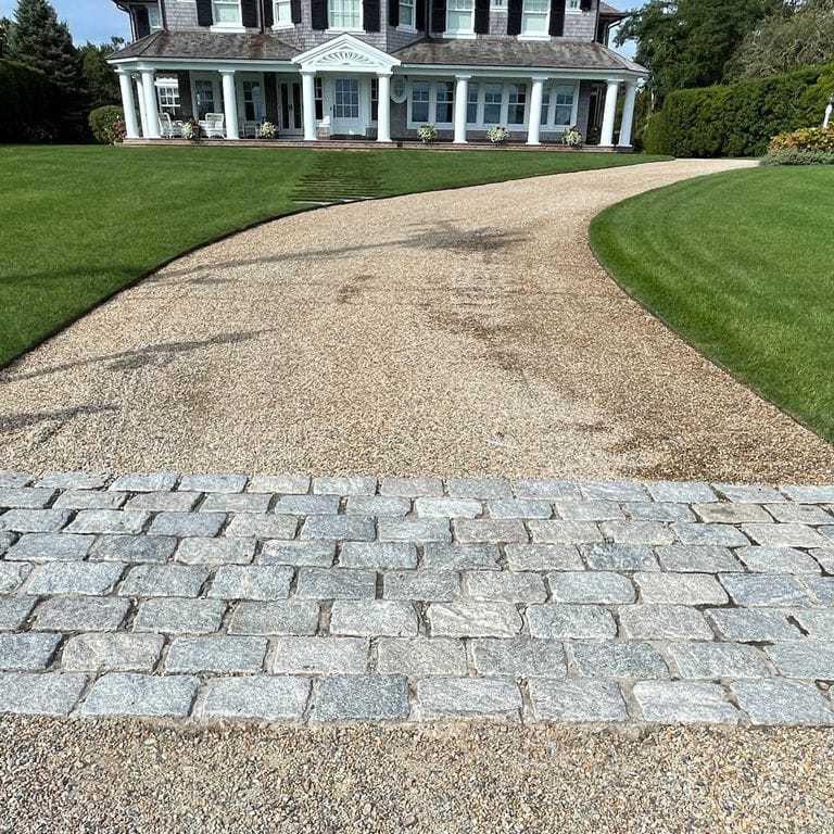 Application image for Granite Cobblestones - Gray. The image shows this material being used in a driveway aprons application. The surface finish shown is a combination of split-face and tumbled. The dominant color shown is gray; with a white secondary. The color mix creates a cool feeling.