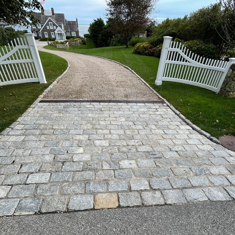 Application image for Granite Cobblestones - Gray. The image shows this material being used in a driveway aprons application. The surface finish shown is tumbled. The dominant color shown is gray; with white and tan secondaries. Exhibits "movement" and the color mix creates a cool feeling. Marbled and speckled color patterns.