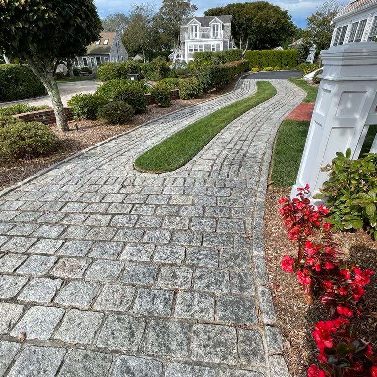 Application image for Granite Cobblestones - Gray. The image shows this material being used in a driveway pavers application. The surface finish shown is tumbled. The dominant color shown is gray; with a white secondary. The color mix creates a cool feeling. Blotched color pattern.