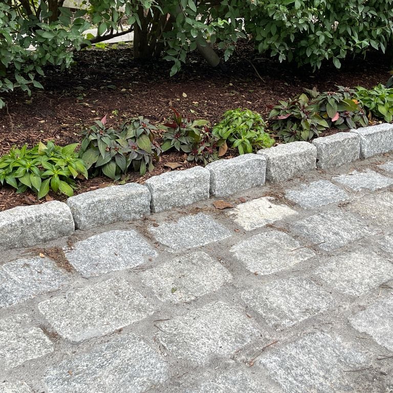 Application image for Granite Cobblestones - Gray. The image shows this material being used for landscape curbing &amp; edging and driveway pavers. The surface finish shown is tumbled. The dominant color shown is gray; with white and cream secondaries. The color mix creates a cool feeling.