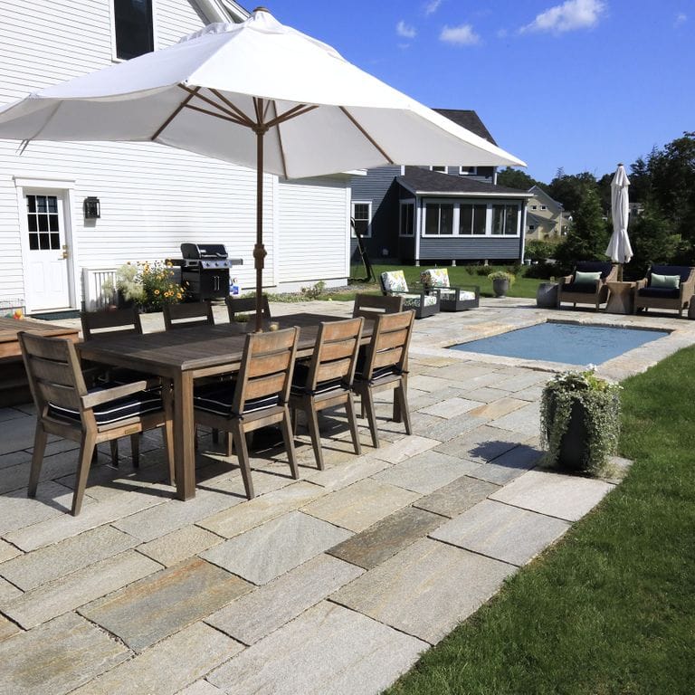 Feature image for Pietra Italia Full Color Gneiss. The image shows this material being used for patio pavers and pool deck pavers. The surface finish shown is natural cleft. Dominant colors shown are beige, gray and tan; with brown, orange/rust, purple, white and cream secondaries. The color mix creates a warm feeling. Regular and random color patterns. Contains some mica and mineral inclusions.