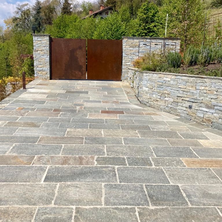 Application image for Pietra Italia Full Color Gneiss. The image shows this material being used for landscape wallstone and driveway pavers. The surface finish shown is natural cleft. Dominant colors shown are beige and gray; with beige, orange/rust, white, tan and cream secondaries. The color mix creates a warm feeling. Regular and fine speckled color patterns.