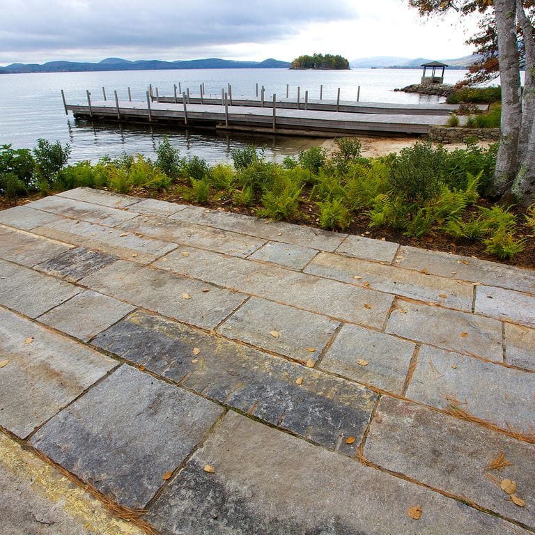 Feature image for Reclaimed First Generation Granite Curbstone. The image shows this material being used in a patio pavers application. Design patterns shown are roughly square roughly rectangular, planks with constant or same size and planks with varied heights and lengths. The dominant color shown is gray; with beige, brown, orange/rust, purple, white and tan secondaries. The color mix creates a warm feeling. Regular, speckled and blotched color patterns.