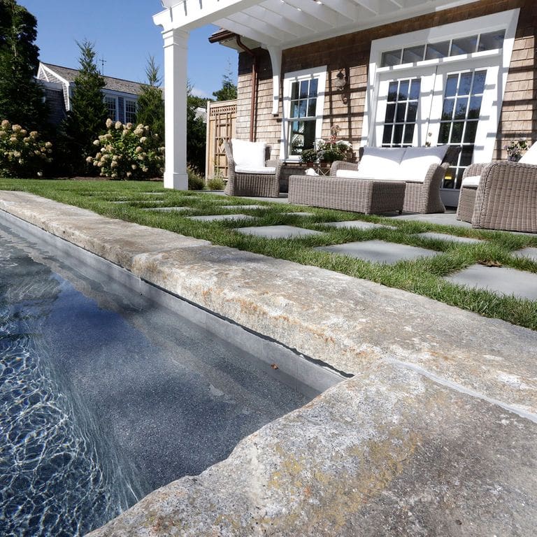 Application image for Reclaimed First Generation Granite Curbstone. The image shows this material being used in a pool coping application. The surface finish shown is a combination of split-face with flat plane, weathered and worn. The dominant color shown is gray; with beige, brown, orange/rust, white, tan and cream secondaries. The color mix creates a warm feeling. Regular and speckled color patterns.