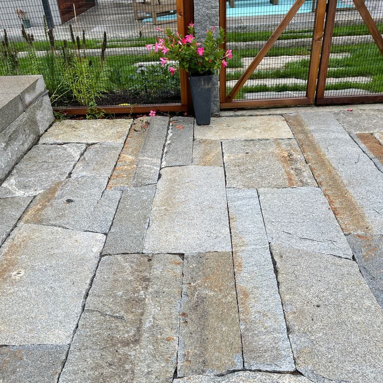 Application image for Reclaimed Second Generation Granite Curbstone. The image shows this material being used for patio pavers and pool deck pavers. The surface finish shown is a combination of split-face with flat plane, weathered and worn. The dominant color shown is gray; with brown, orange/rust and tan secondaries. Exhibits "movement" and the color mix creates a warm feeling. Linear, speckled and blotched color patterns.