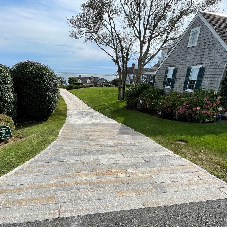 Application image for Reclaimed Third Generation Granite Curbstone. The image shows this material being used in a driveway aprons application. The surface finish shown is a combination of weathered and worn. The dominant color shown is gray; with a beige secondary. Exhibits "movement". Linear and speckled color patterns.