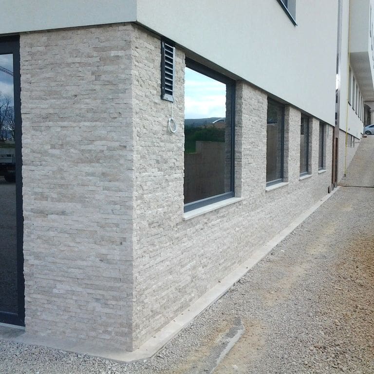 Application image for Dalmatian Limestone. The image shows this material being used for full bed veneer and thin veneer. The surface finish shown is split-face with flat plane. Dominant colors shown are beige, gray and white; with pink, purple and cream secondaries. The color mix creates a cool feeling. Regular color pattern.