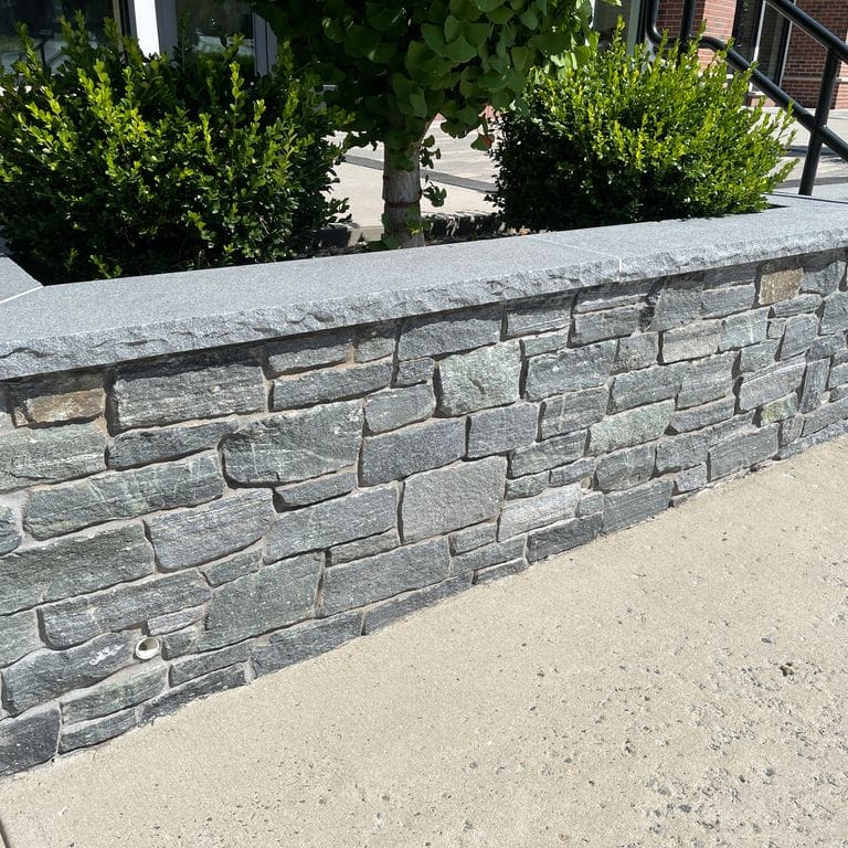 Feature image for Connecticut White Line Granite. The image shows this material being used in a landscape wallstone application. The surface finish shown is split-face. Dominant colors shown are gray and white; with green and white secondaries. Exhibits "movement" and the color mix creates a cool feeling. Regular and linear color patterns.