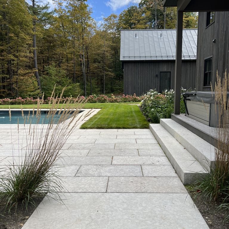 Feature image for Limestone - Madison. The image shows this material being used for pool coping, path pavers, patio pavers and monolithic steps. The surface finish shown is flamed. The dominant color shown is gray; with beige, blue, purple, white and cream secondaries. The color mix creates a cool feeling. Regular and cloudy color patterns.