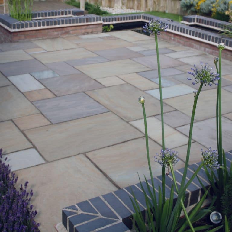 Application image for Lakeland Sandstone. The image shows this material being used in a patio pavers application. The surface finish shown is natural cleft. Dominant colors shown are blue and gray; with green and white secondaries. Regular and streaked color patterns.