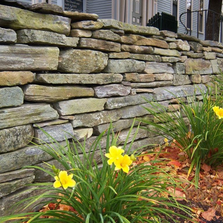 Feature image for Mystic Mountain Natural - Wallstone. The image shows this product being used in a landscape wallstone application. The design pattern shown is ledgestone. Dominant colors shown are brown, green and gray; with orange/rust, purple and tan secondaries. Exhibits "movement" and the color mix creates a warm feeling. Random, speckled and blotched color patterns.