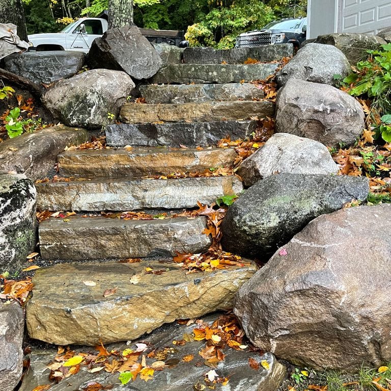 Application image for New England Fieldstone - Boulders. The image shows this product being used in a landscape boulders &amp; ledge application. The surface finish shown is weathered. The dominant color shown is gray; with beige, brown, white and tan secondaries. The color mix creates a warm feeling. Regular color pattern.